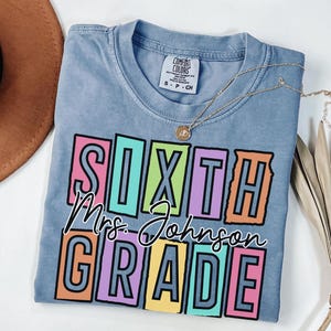 May include: A light blue Comfort Colors t-shirt with the words "SIXTH Mrs. Johnson GRADE" in a colorful block letter design. A gold necklace with a pendant is draped across the shirt.