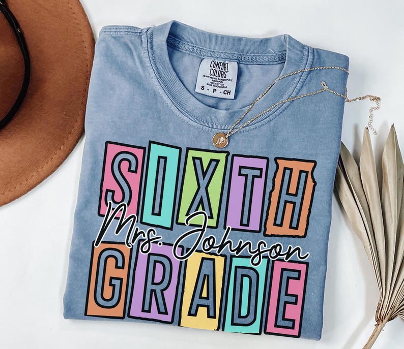 May include: A light blue Comfort Colors t-shirt with the words "SIXTH Mrs. Johnson GRADE" in a colorful block letter design. A gold necklace with a pendant is draped across the shirt.