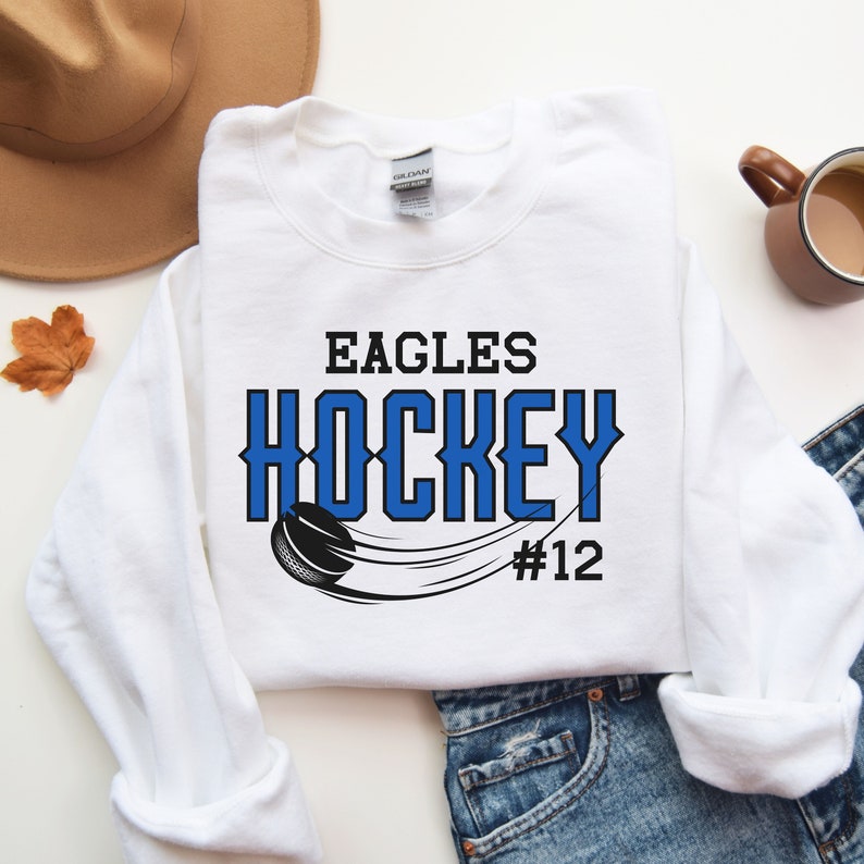 K&ouml;nnte beinhalten: Wei&szlig;er Pullover mit blauem und schwarzem Text, der "Eagles Hockey #12" lautet, mit einer Grafik von einem Hockeypuck und einem Schl&auml;ger.