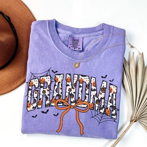 May include: A lilac Comfort Colors t-shirt with the word "GRANDMA" in a Halloween-themed design. The letters are filled with pumpkins and bats, with spiderwebs and an orange bow. A gold necklace and a brown hat are also in the image.