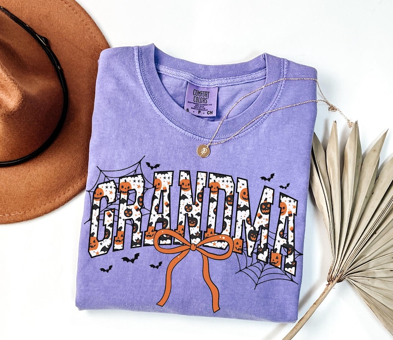 May include: A lilac Comfort Colors t-shirt with the word "GRANDMA" in a Halloween-themed design. The letters are filled with pumpkins and bats, with spiderwebs and an orange bow. A gold necklace and a brown hat are also in the image.