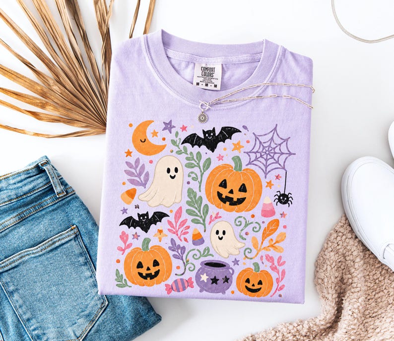 Może przedstawiać: Jasnofioletowy t-shirt z kolorowym nadrukiem graficznym o tematyce Halloween. Wz&oacute;r zawiera pomarańczowe dynie, białe duszki, czarne nietoperze, pajęczynę z pająkiem, ż&oacute;łty p&oacute;łksiężyc, cukierki w kształcie kukurydzy oraz fioletowy kociołek.