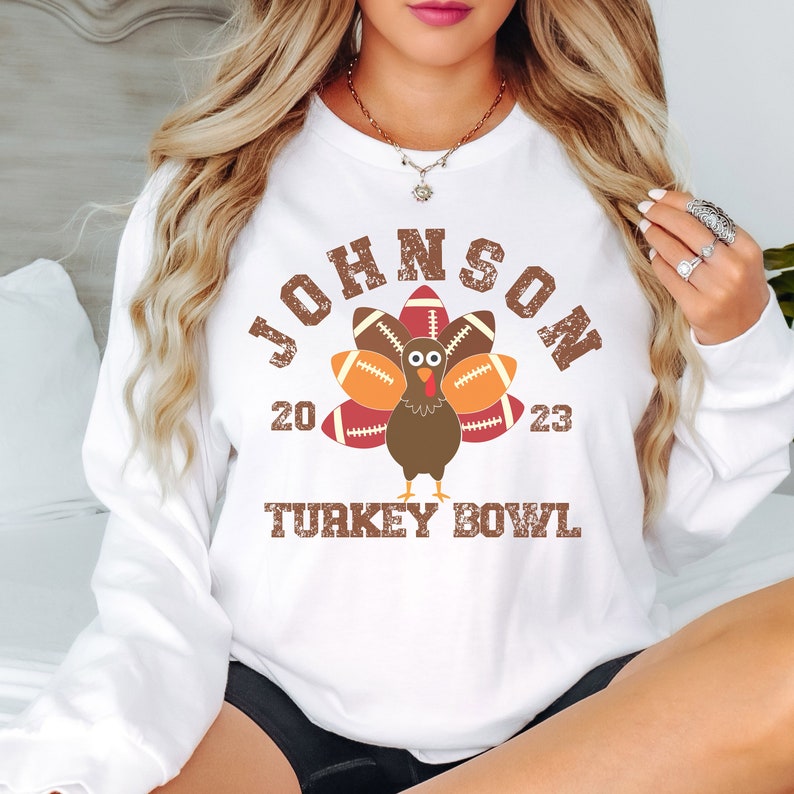 Op de afbeelding: Wit sweatshirt met een bruine kalkoen die voetballen als veren draagt. De tekst "JOHNSON 20 23 TURKEY BOWL" staat op het sweatshirt gedrukt.