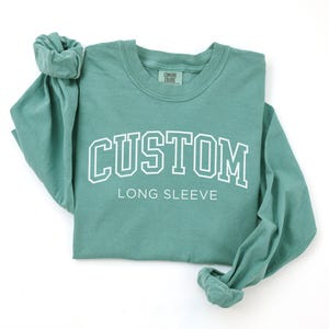 Op de afbeelding: Een tealkleurig shirt met lange mouwen met het woord "CUSTOM" in witte blokletters boven "LONG SLEEVE". Het shirt is gemaakt van een zacht materiaal en heeft een ronde hals. De mouwen zijn opgerold.