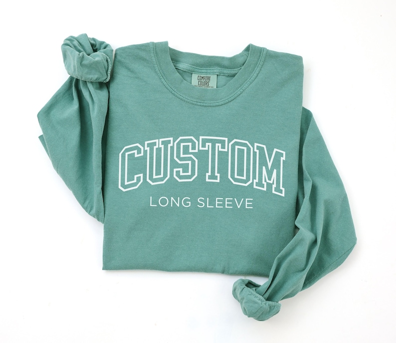 Op de afbeelding: Een tealkleurig shirt met lange mouwen met het woord "CUSTOM" in witte blokletters boven "LONG SLEEVE". Het shirt is gemaakt van een zacht materiaal en heeft een ronde hals. De mouwen zijn opgerold.