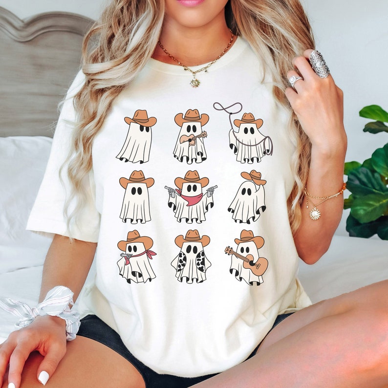 May include: White t-shirt with a graphic of nine cartoon ghosts wearing cowboy hats and doing various activities. The ghosts are wearing bandanas, playing a guitar, holding a lasso, and holding a gun.