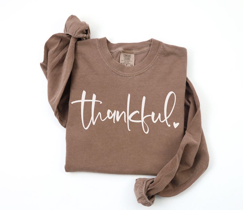 May include: A brown long-sleeve shirt with the word "thankful" in white script. The shirt has a crew neck and a small heart at the end of the word. The shirt is made of a soft material.
