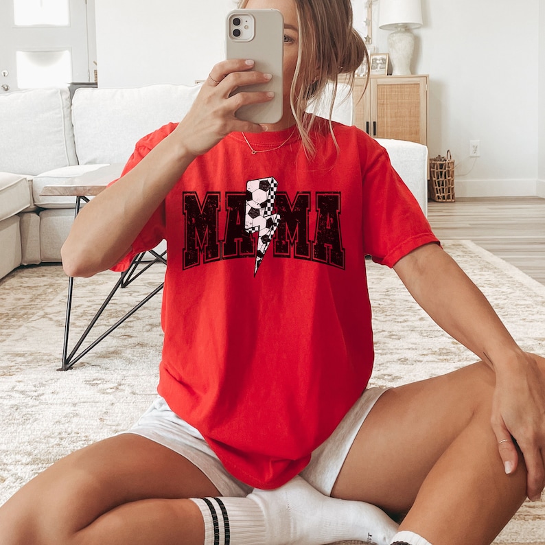May include: Red t-shirt with a black "MAMA" graphic featuring a lightning bolt design with a soccer ball pattern.