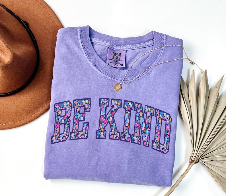 May include: A lavender Comfort Colors t-shirt with the words "BE KIND" in a floral design. A gold necklace with a pendant is draped across the shirt. A brown hat with a black band is in the background.