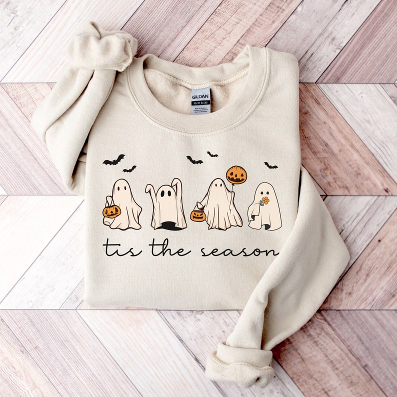 May include: Beige sweatshirt with a Halloween-themed graphic. The design features four cartoon ghosts, bats, and pumpkins. The text "tis the season" is printed below the ghosts. The sweatshirt is folded on a wooden surface.
