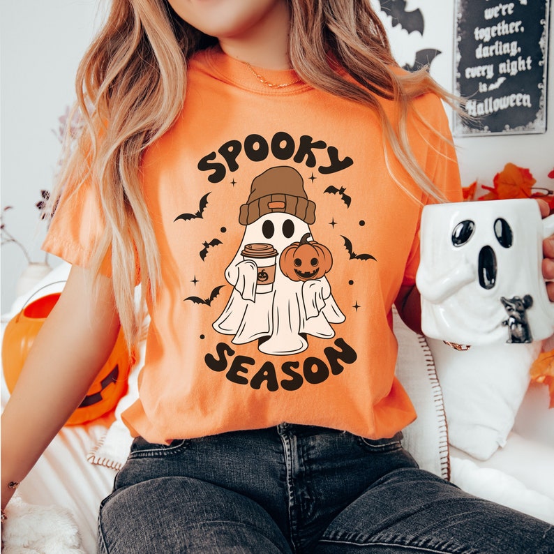 Op de afbeelding: Oranje T-shirt met een cartoon-spook dat een pompoen en een kopje koffie vasthoudt. De tekst "Spooky Season" staat op het shirt gedrukt.