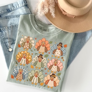May include: A sage green t-shirt featuring a graphic print of nine turkeys with various patterned feathers in shades of orange, blue, pink, and brown, interspersed with small leaves and stars, accompanied by light blue denim jeans and a tan hat.