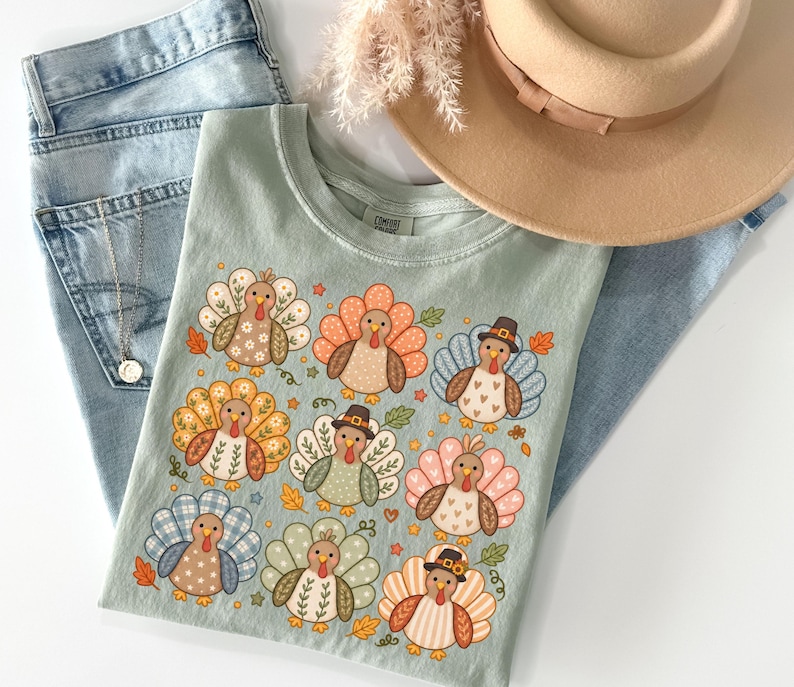 May include: A sage green t-shirt featuring a graphic print of nine turkeys with various patterned feathers in shades of orange, blue, pink, and brown, interspersed with small leaves and stars, accompanied by light blue denim jeans and a tan hat.