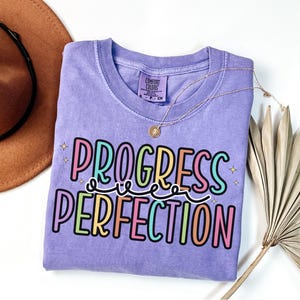 May include: A lilac Comfort Colors t-shirt with the words "PROGRESS PERFECTION" in a colorful, playful font. The shirt is adorned with a gold necklace and a decorative palm leaf. A brown hat is visible in the background.
