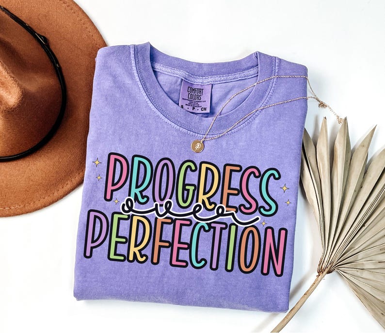 May include: A lilac Comfort Colors t-shirt with the words "PROGRESS PERFECTION" in a colorful, playful font. The shirt is adorned with a gold necklace and a decorative palm leaf. A brown hat is visible in the background.