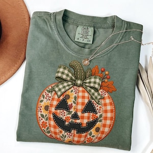 May include: A sage green t-shirt featuring a large pumpkin graphic with a black jack-o'-lantern face. The pumpkin has orange gingham and floral patterned sections, topped with a green and white gingham bow, orange leaves, and berries.