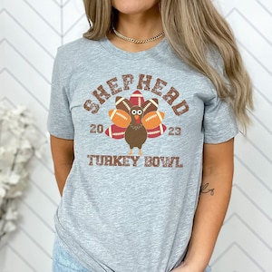 Op de afbeelding: Een grijs T-shirt met een verweerde afbeelding van een kalkoen die een voetbalhelm draagt en voetballen vasthoudt. De tekst "SHEPHERD 20 23 TURKEY BOWL" is op het T-shirt gedrukt.