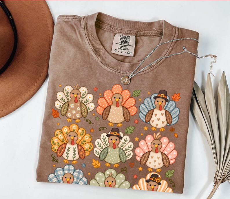 May include: A brown short-sleeved t-shirt with a Comfort Colors label, featuring a repeating graphic print of nine stylized turkeys in various patterns and colors, including some wearing hats, surrounded by small autumn leaves and floral accents.