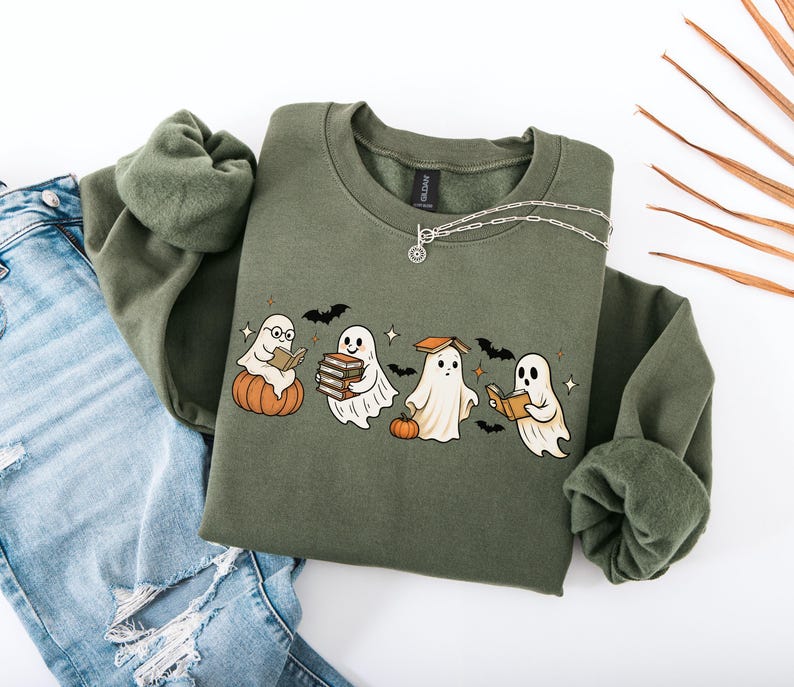 Puede incluir: Una sudadera verde oliva con un gr&aacute;fico de cuatro fantasmas de dibujos animados leyendo libros, con calabazas y murci&eacute;lagos. Un collar plateado est&aacute; drapeado sobre la sudadera. La sudadera se combina con unos vaqueros azules.