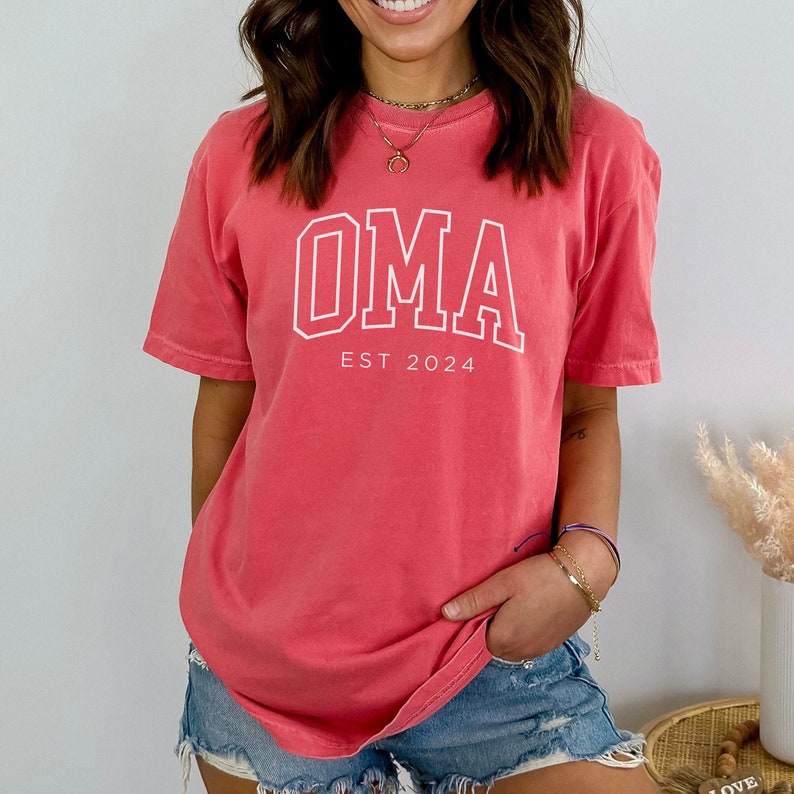 Pu&ograve; includere: Una t-shirt color corallo con una grafica bianca che dice "OMA EST 2024".