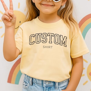 Puede incluir: Una camiseta de manga corta de color amarillo claro que presenta la palabra CUSTOM en una fuente negra en negrita de estilo universitario, con la palabra SHIRT impresa en una fuente negra más pequeña y sencilla en el área del pecho.