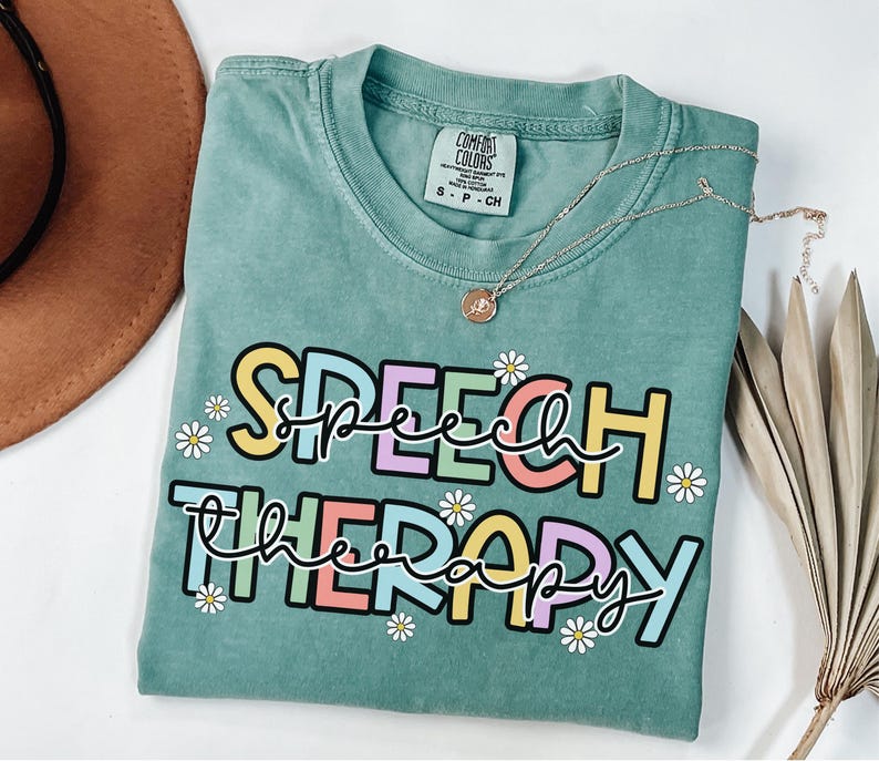 May include: A teal Comfort Colors t-shirt with the words "SPEECH THERAPY" in colorful, bubble letters and daisy accents. A gold necklace with a pendant rests on the shirt. The shirt is folded and placed on a white surface.