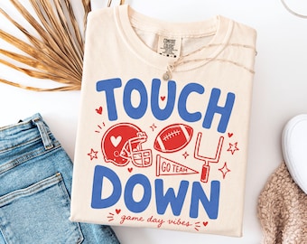 Puede incluir: Una camiseta de color crema con el texto azul TOUCH DOWN y gráficos rojos de un casco de fútbol americano, un balón, un poste de meta y un banderín que dice GO TEAM. La camiseta también incluye el texto rojo game day vibes y corazones.