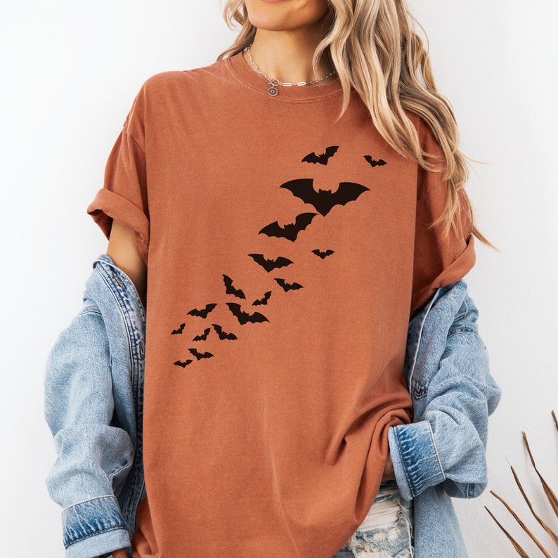 Puede incluir: Una camiseta de color &oacute;xido con un gr&aacute;fico de una bandada de murci&eacute;lagos volando en diagonal. Los murci&eacute;lagos son negros y est&aacute;n impresos en la parte delantera de la camiseta.