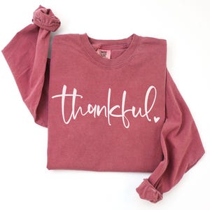 May include: A long-sleeved, maroon-colored shirt with the word "thankful" in white script across the front. A small white heart is next to the word. The sleeves are cuffed at the wrists. The shirt is on a white background.