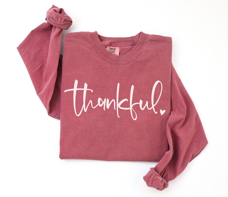 May include: A long-sleeved, maroon-colored shirt with the word "thankful" in white script across the front. A small white heart is next to the word. The sleeves are cuffed at the wrists. The shirt is on a white background.