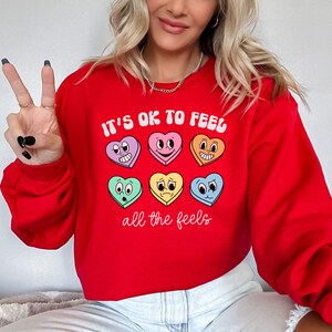 Pu&ograve; includere: Felpa rossa con il testo "It's OK to feel all the feels" e una fila di caramelle colorate a forma di cuore con diverse espressioni facciali.