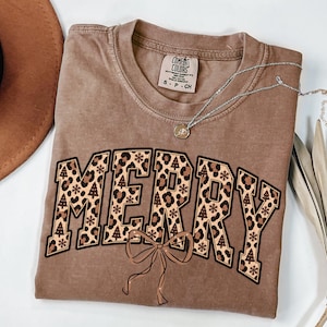 Puede incluir: Una camiseta marrón de manga corta con cuello redondo que presenta la palabra MERRY en letras grandes. Las letras tienen un estampado de leopardo con pequeños iconos de árboles de Navidad y copos de nieve. Un lazo está impreso bajo el texto.