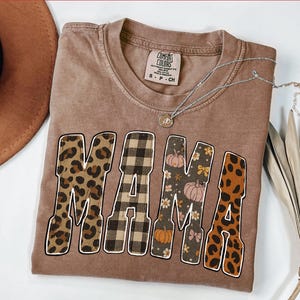 May include: A brown Comfort Colors t-shirt with the word "MAMA" in a mix of leopard print, plaid, floral, and pumpkin designs. A silver necklace with a small pendant is draped across the shirt.