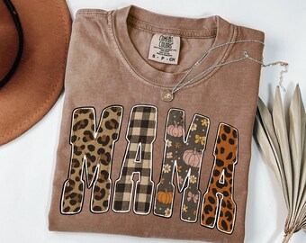 May include: A brown Comfort Colors t-shirt with the word "MAMA" in a mix of leopard print, plaid, floral, and pumpkin designs. A silver necklace with a small pendant is draped across the shirt.