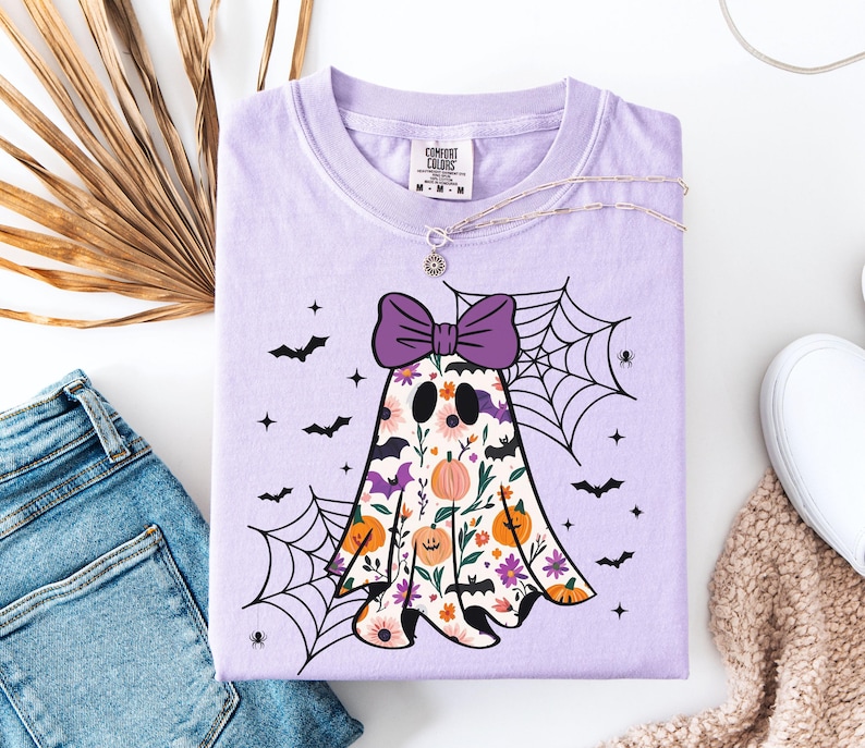 May include: A lilac Comfort Colors t-shirt with a floral ghost design, a purple bow, bats, and spiderwebs. The ghost has a floral pattern with pumpkins and flowers. A silver necklace is on the shirt.