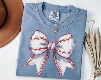 May include: A light blue t-shirt with a white and red baseball-themed bow. The shirt has the text "COMFORT COLORS" on the tag.