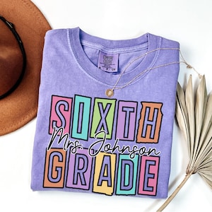 May include: A light purple Comfort Colors t-shirt with the words "SIXTH GRADE Mrs. Johnson" in a colorful block design. A gold necklace and a brown hat are visible in the background. The shirt is a crew neck and has a small tag on the collar.