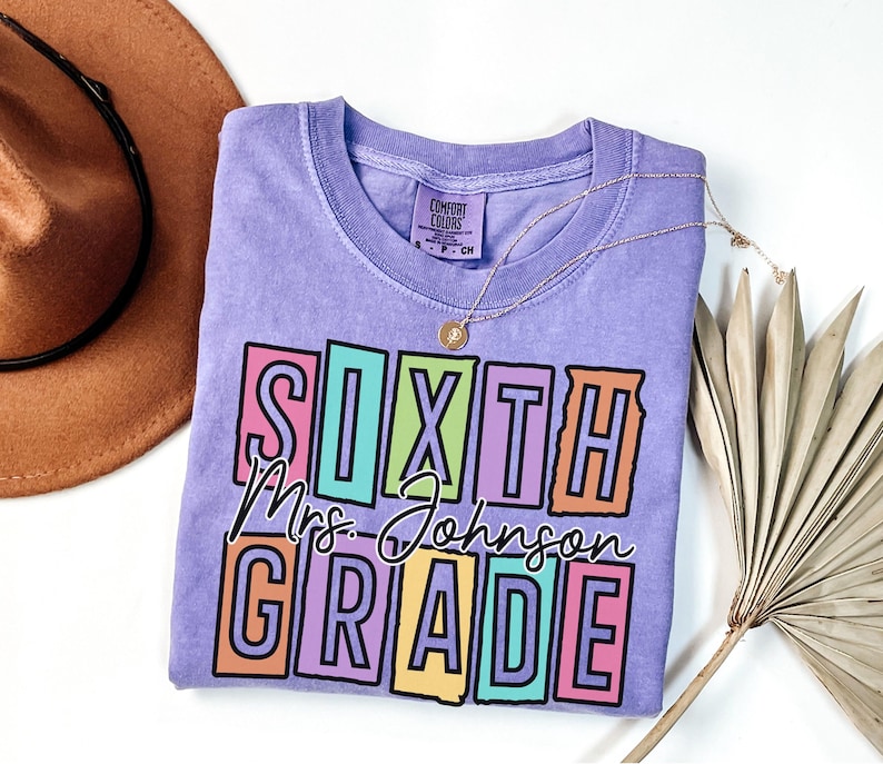 May include: A light purple Comfort Colors t-shirt with the words "SIXTH GRADE Mrs. Johnson" in a colorful block design. A gold necklace and a brown hat are visible in the background. The shirt is a crew neck and has a small tag on the collar.