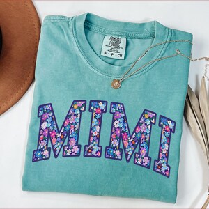 Puede incluir: Una camiseta turquesa Comfort Colors con la palabra "MIMI" en un dise&ntilde;o floral. La camiseta tiene cuello redondo y mangas cortas. Un collar dorado con un colgante est&aacute; sobre la camiseta.