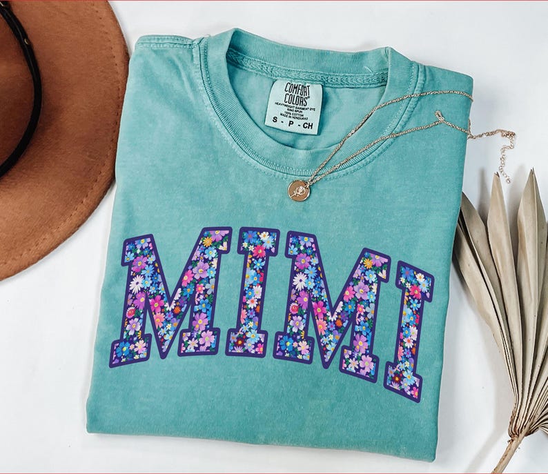 Puede incluir: Una camiseta turquesa Comfort Colors con la palabra "MIMI" en un dise&ntilde;o floral. La camiseta tiene cuello redondo y mangas cortas. Un collar dorado con un colgante est&aacute; sobre la camiseta.