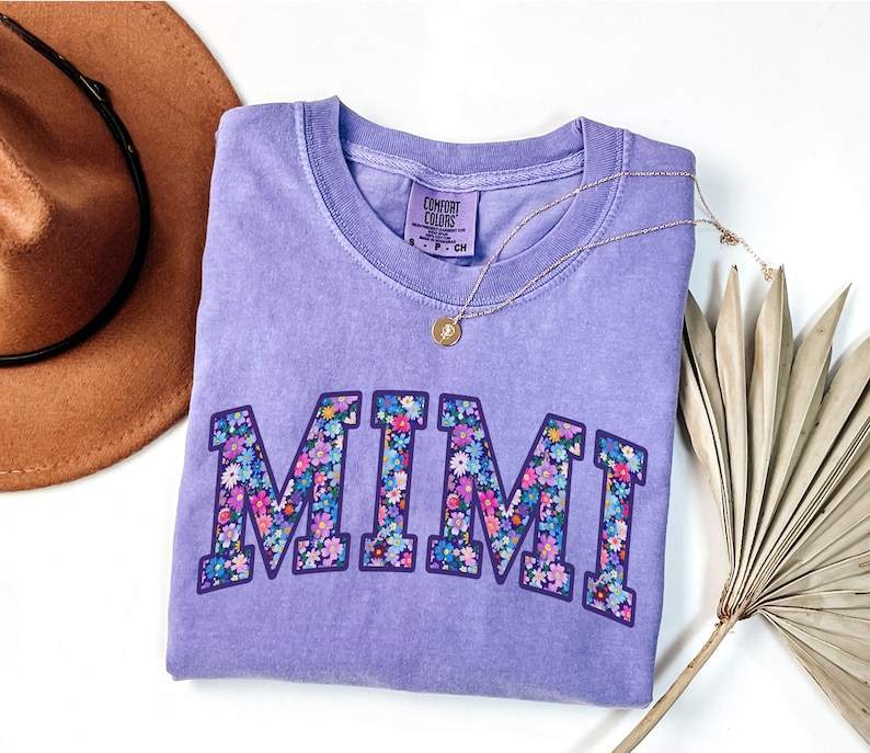 Puede incluir: Una camiseta Comfort Colors de color lila con la palabra "MIMI" en un dise&ntilde;o floral. Un collar de oro con un colgante est&aacute; drapeado sobre la camisa. Un sombrero marr&oacute;n y una hoja de palma seca est&aacute;n en el fondo.