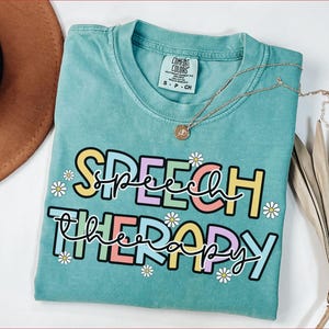 May include: A turquoise Comfort Colors t-shirt with the words "SPEECH THERAPY" in a colorful, whimsical font. The design includes daisy accents. A gold necklace with a pendant is draped across the shirt.
