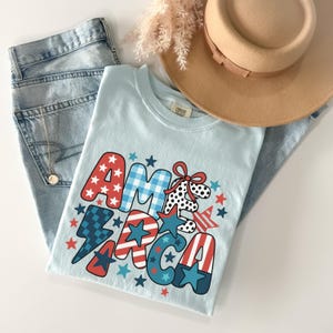 May include: A light blue t-shirt with the word "AMERICA" in a patriotic design, featuring red, white, and blue stars and patterns. The shirt is paired with light wash denim jeans, a tan hat, and a gold necklace. A casual, patriotic theme.