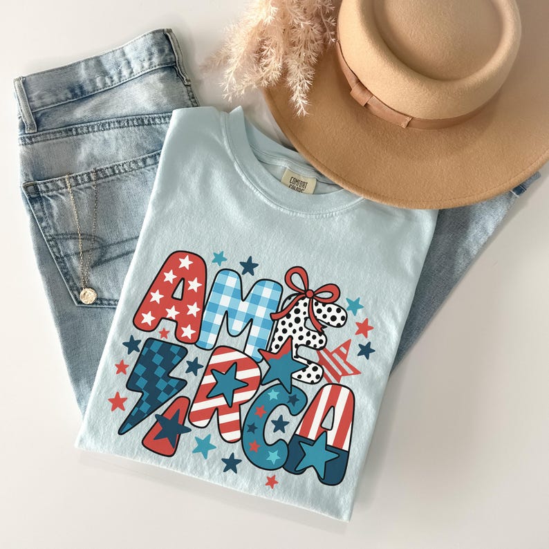 May include: A light blue t-shirt with the word "AMERICA" in a patriotic design, featuring red, white, and blue stars and patterns. The shirt is paired with light wash denim jeans, a tan hat, and a gold necklace. A casual, patriotic theme.