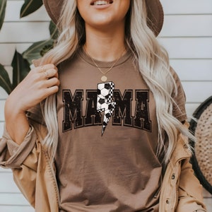 May include: A brown t-shirt with a distressed black graphic that says "MAMA" with a lightning bolt and soccer ball design.