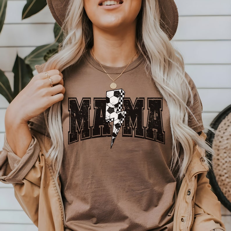 May include: A brown t-shirt with a distressed black graphic that says "MAMA" with a lightning bolt and soccer ball design.