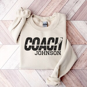 May include: A beige sweatshirt with the text "COACH JOHNSON" in black with a distressed effect. A hockey stick graphic is incorporated into the text.