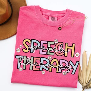 May include: A bright pink t-shirt with the words "SPEECH THERAPY" in a colorful, playful font, decorated with small daisy graphics. A gold necklace with a black pendant rests on the shirt.