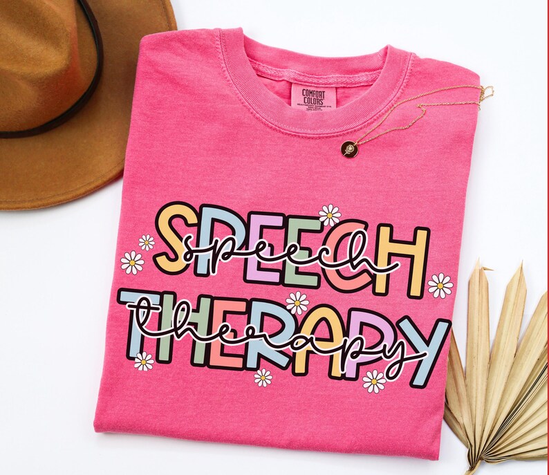 May include: A bright pink t-shirt with the words "SPEECH THERAPY" in a colorful, playful font, decorated with small daisy graphics. A gold necklace with a black pendant rests on the shirt.