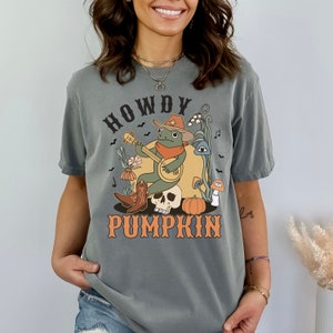 Pu&ograve; includere: Una t-shirt grigio antracite con una stampa grafica di una rana verde che indossa un cappello da cowboy e una bandana, mentre suona il banjo seduta su un teschio. Il disegno include una zucca, funghi, fiori, pipistrelli e il testo HOWDY PUMPKIN.